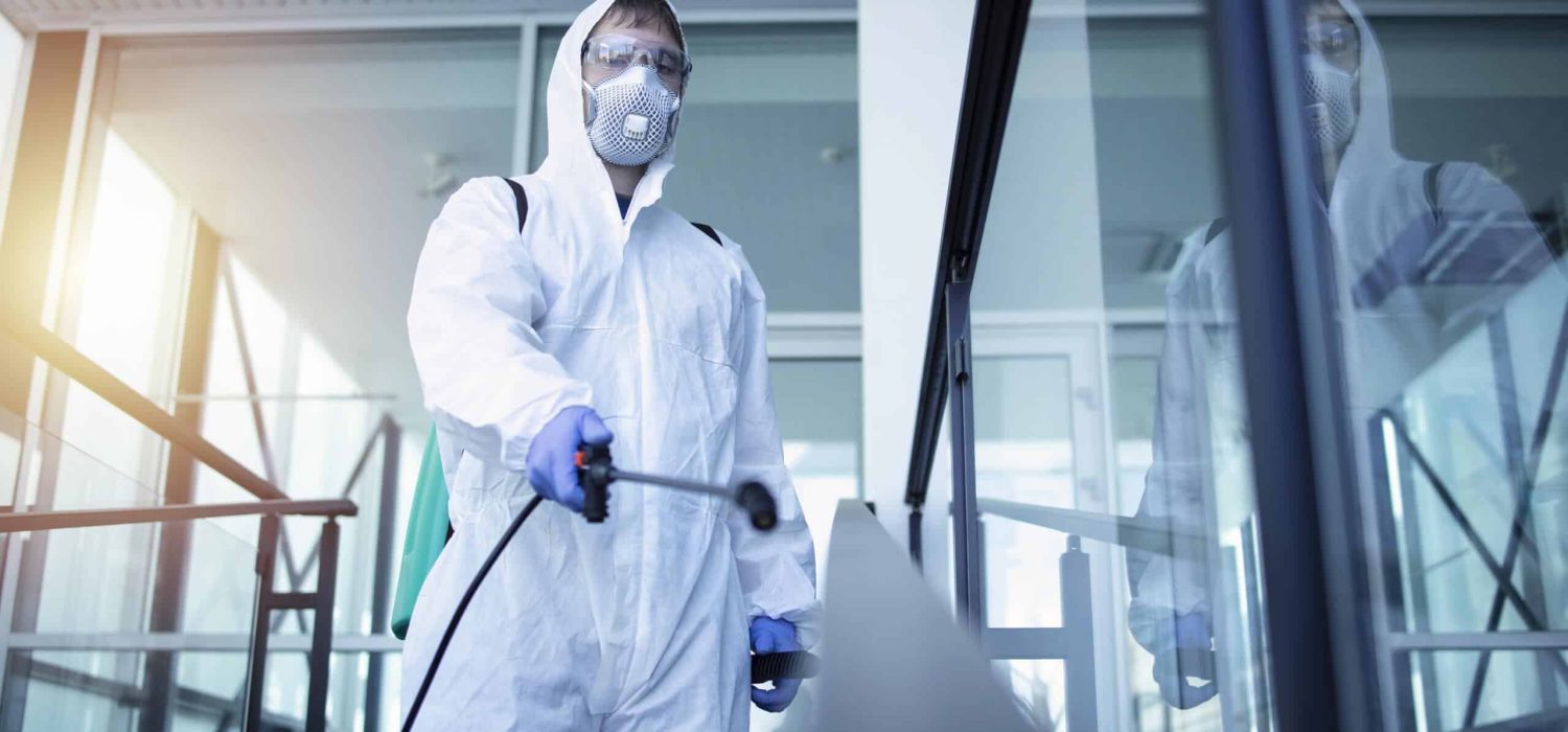 Person in white chemical protection suit doing disinfection of public areas to stop spreading highly contagious corona virus.