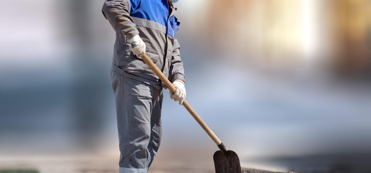 A laborer in a medical mask and work clothes stands with a shovel on a blurry background. Physical labor. Unemployment during the pandemic. Employment and labor.