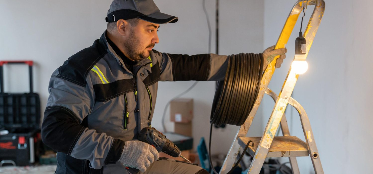 an electrician in overalls with a coil of wires and a drill climbs onto a ladder next to a burning light bulb